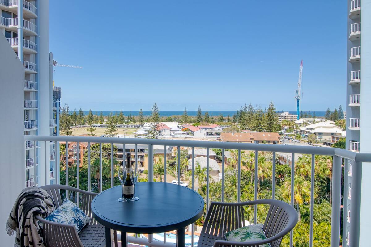 A balcony with a table and chairs, overlooking a coastal town with buildings and a clear sky.