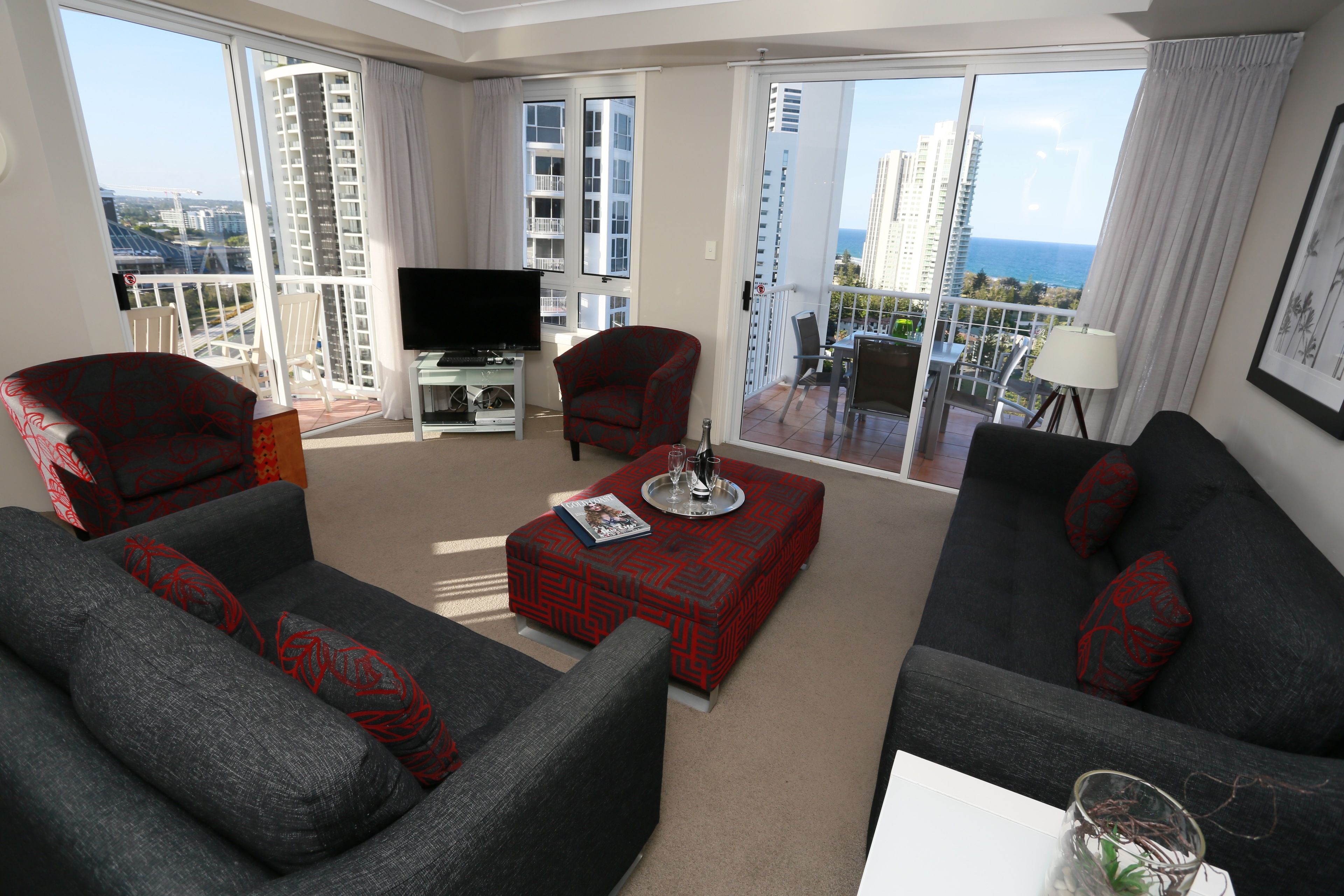 A modern living room with grey sofas, a red patterned ottoman, and a flat-screen TV.