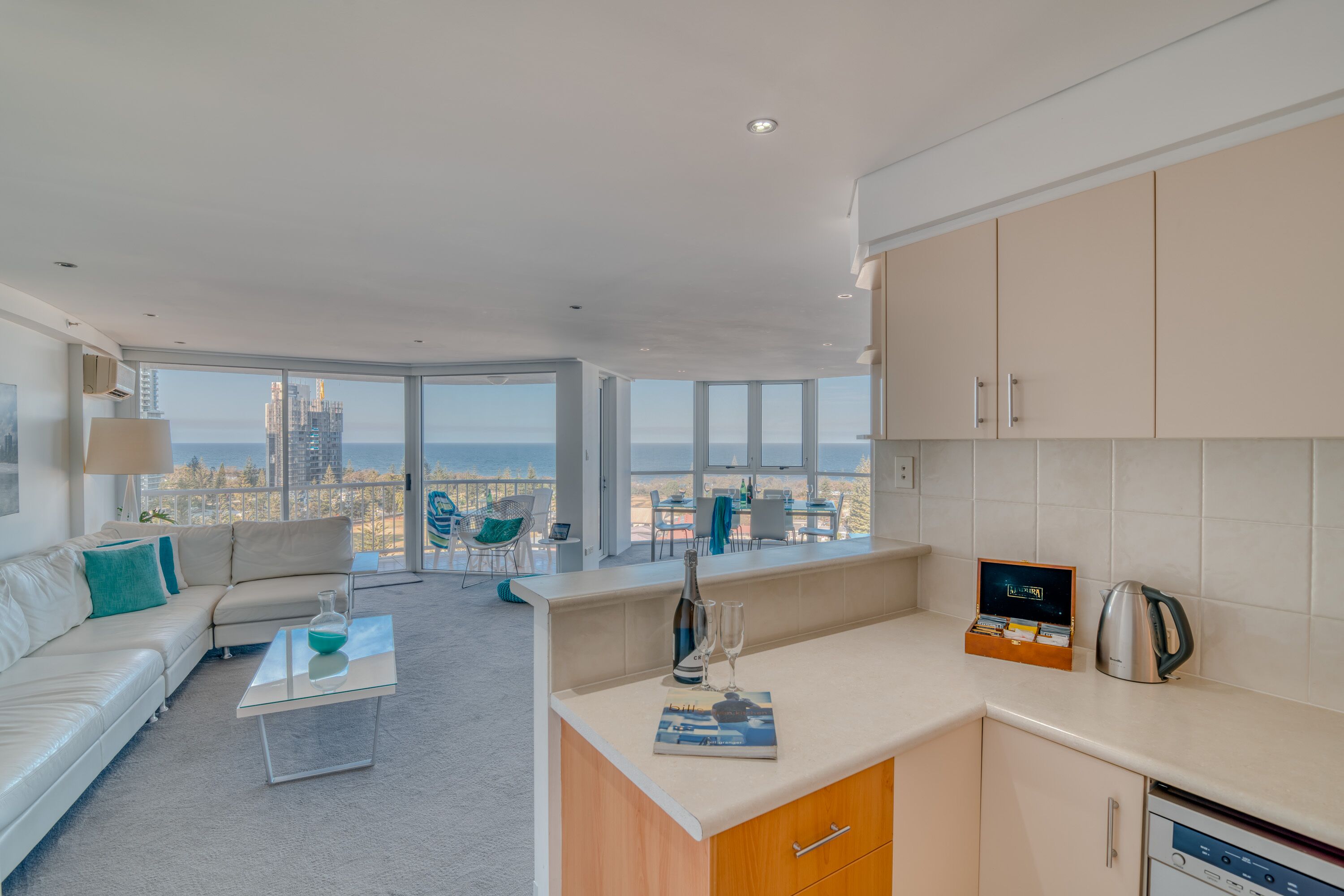 Uma sala de estar moderna com sofá e mesa de centro, área de cozinha com armários e chaleira, e grandes janelas com vista para a cidade.