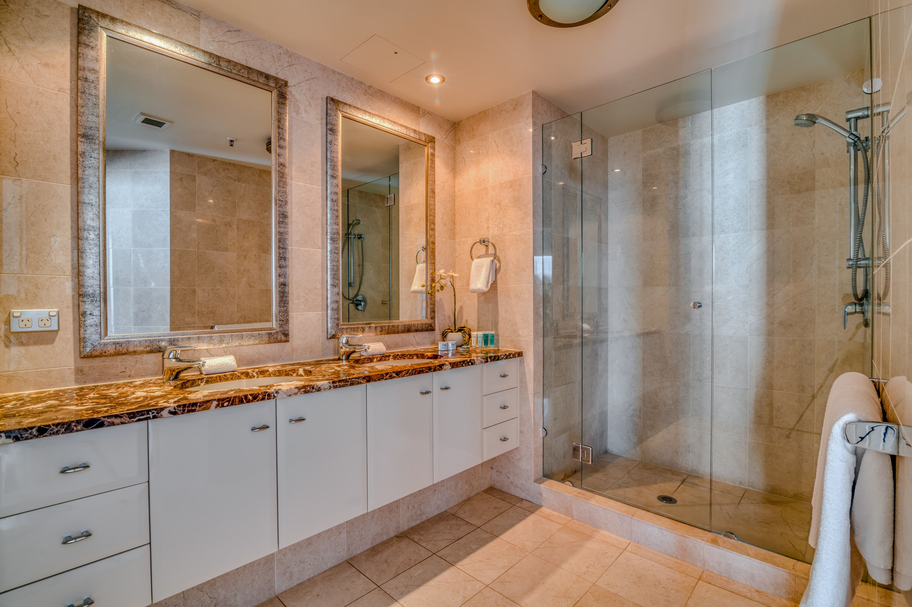 Een moderne badkamer met een marmeren aanrechtblad, twee grote spiegels, een douche met glazen wanden, en een witte handdoek aan de muur.