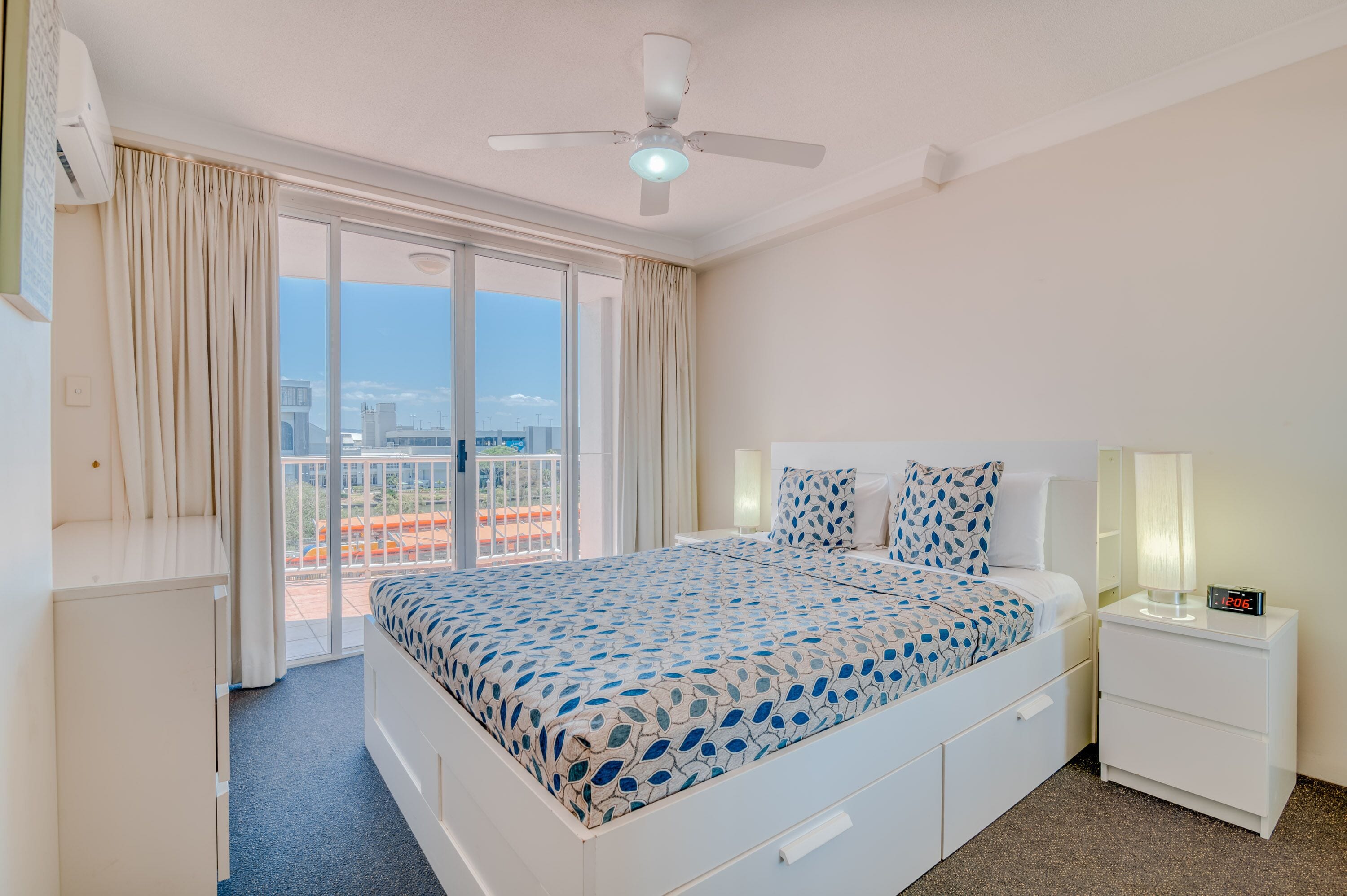 A hotel room with a bed, a nightstand, a ceiling fan, and a view of the city through large windows.