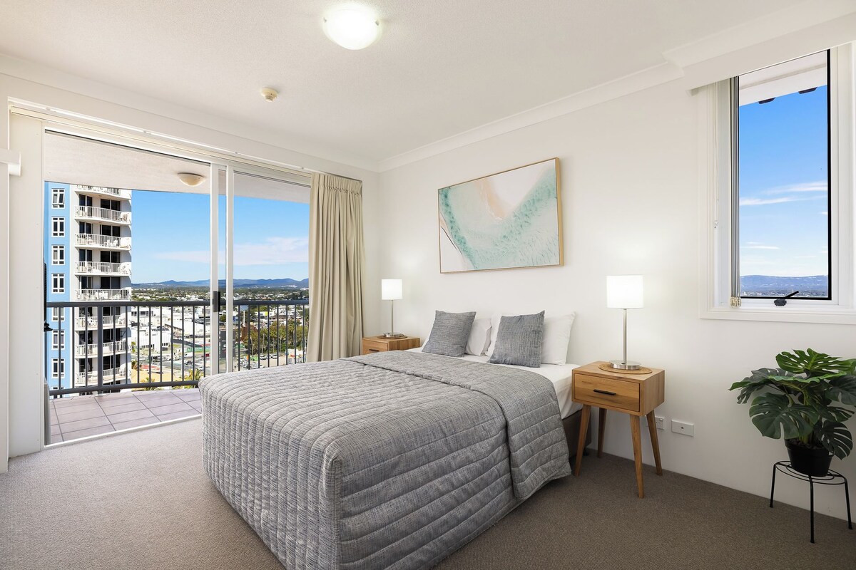 A hotel room with a bed, a nightstand, a lamp, a window with a view, and a plant.