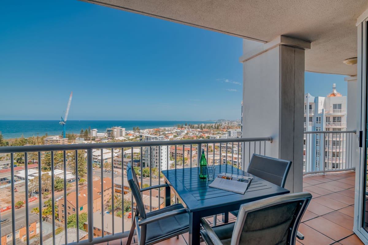 A balcony with a table and chairs, overlooking a coastal cityscape with buildings and a crane.