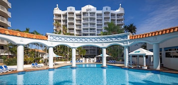 A hotel pool area with a large pool, lounge chairs, and a multi-story building in the background.