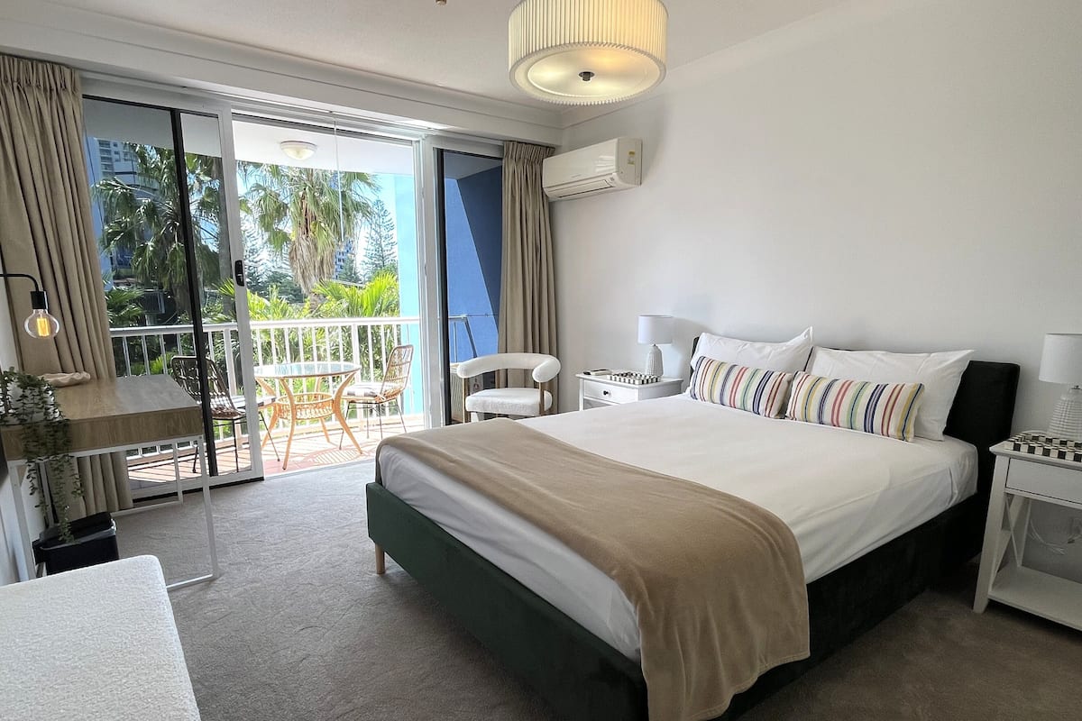 A hotel room with a bed, a desk, a chair, and a balcony with a view of palm trees.