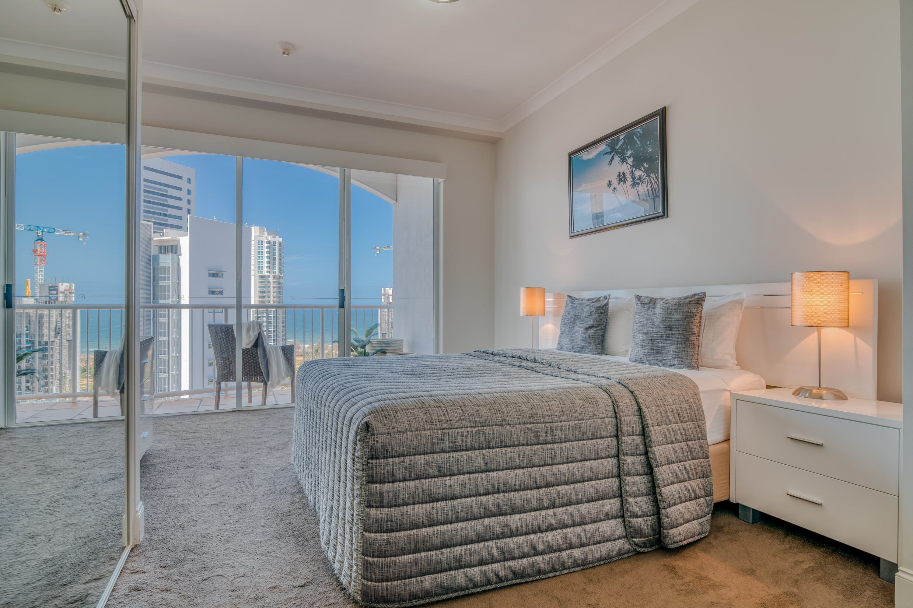 A modern bedroom with a large bed, bedside lamps, a view of the city through floor-to-ceiling windows, and a balcony with outdoor furniture.