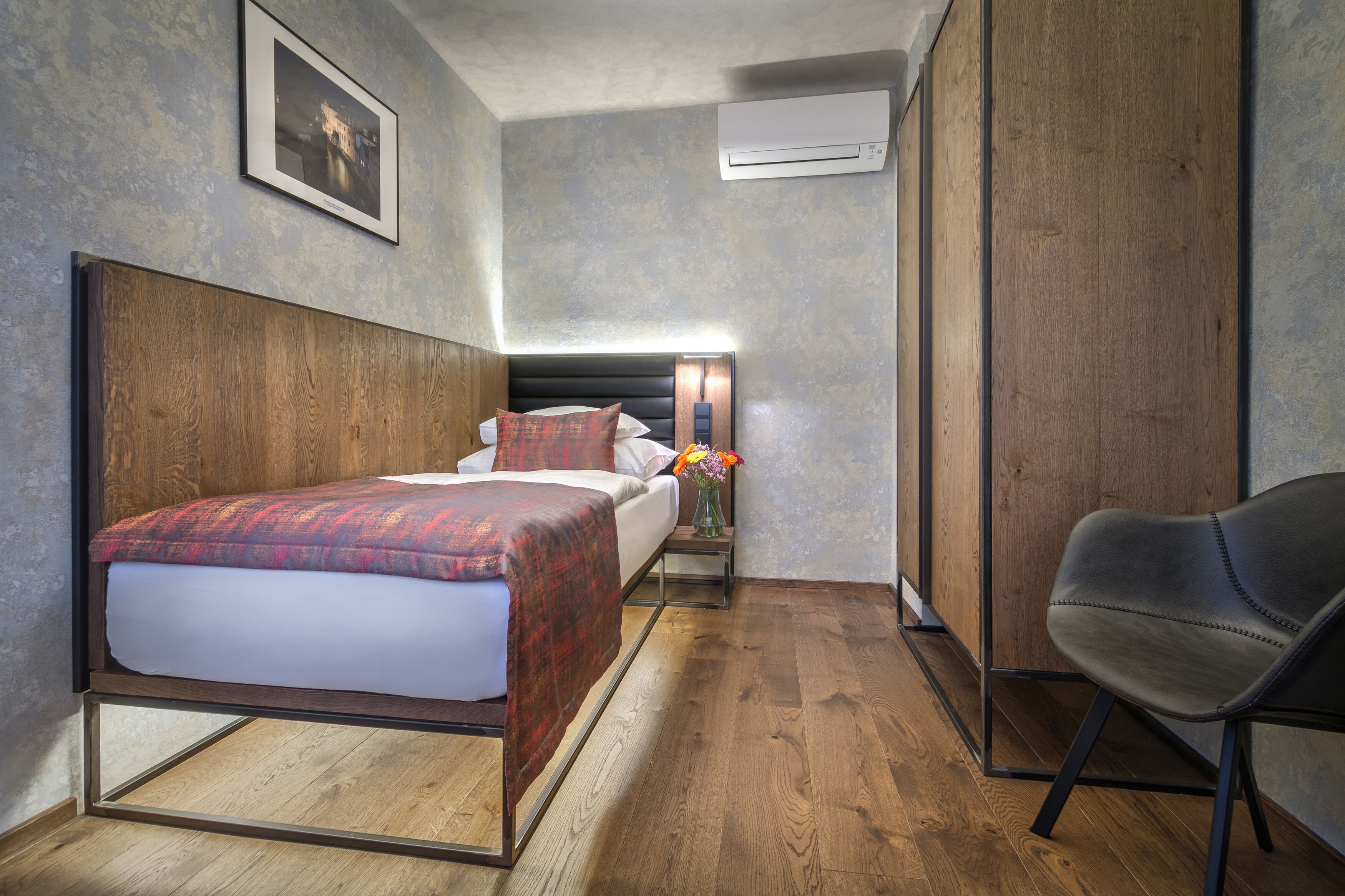 A hotel room with a bed, a chair, an air conditioning unit, and a framed picture on the wall.