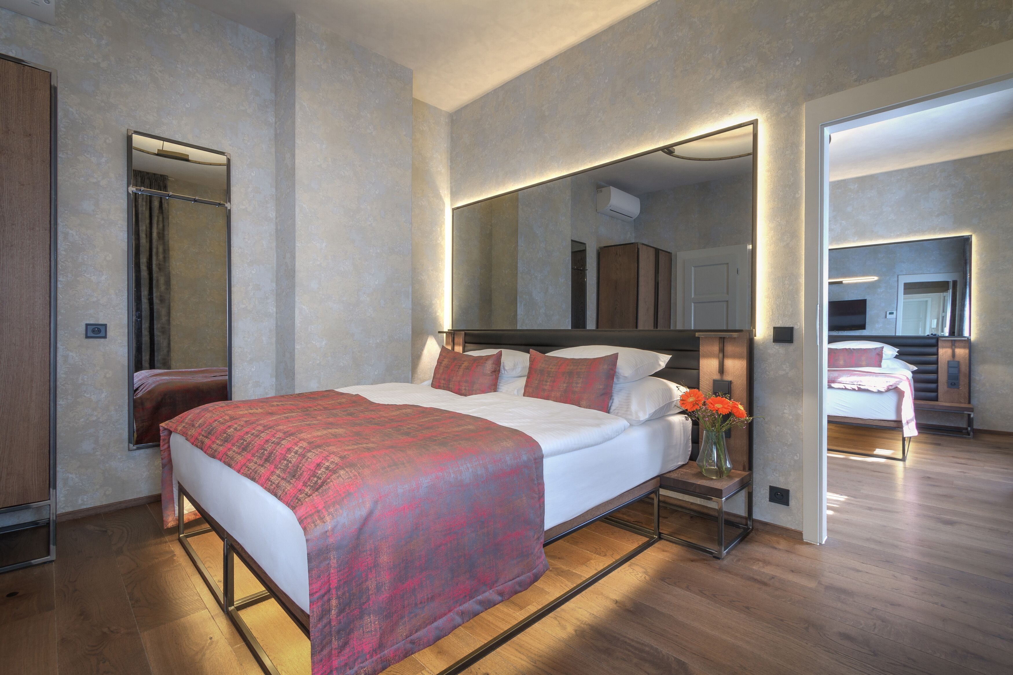 A modern bedroom with a large bed, a bedside table with a vase of flowers, and a mirror above the bed.