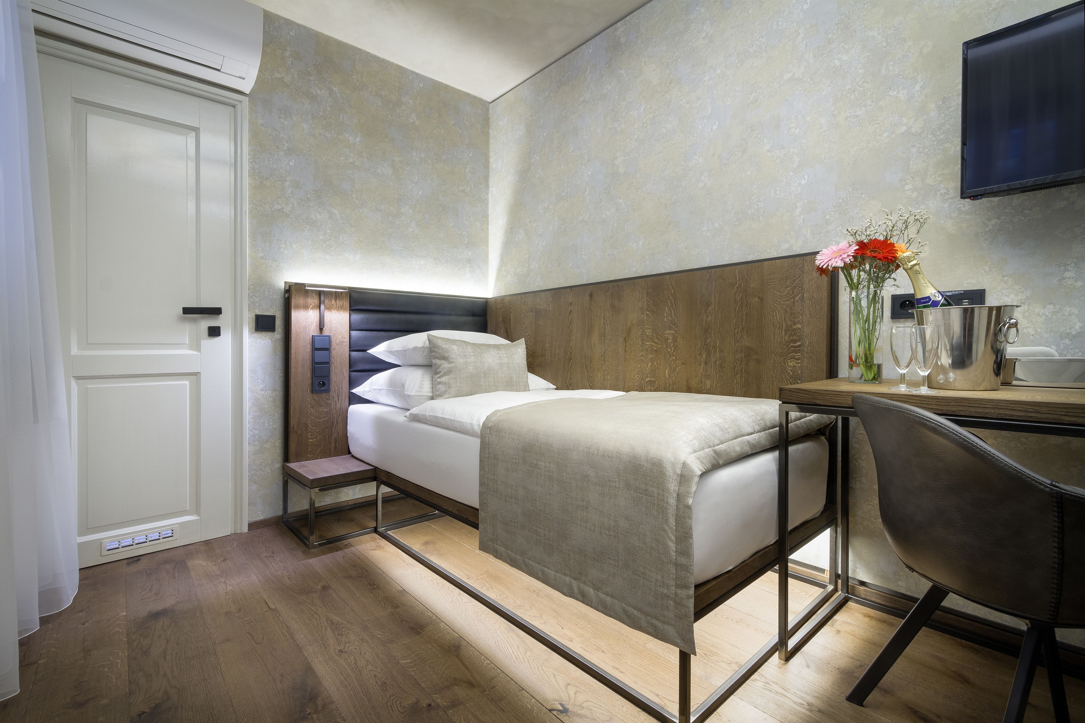 A modern hotel room with a bed, a desk, a chair, and a TV on the wall.