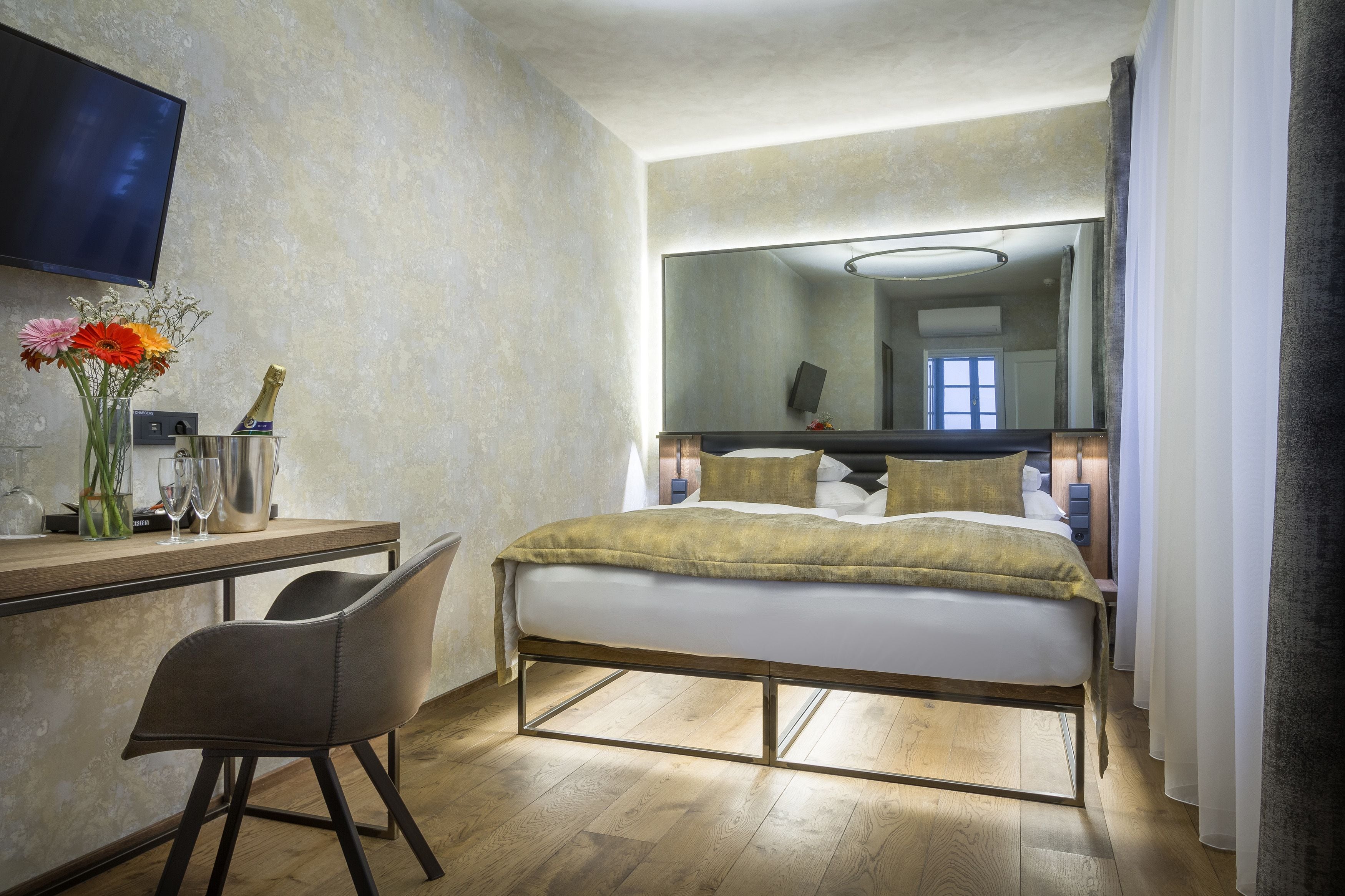 A modern hotel room with a bed, a desk with a chair, a large mirror, and a TV on the wall.