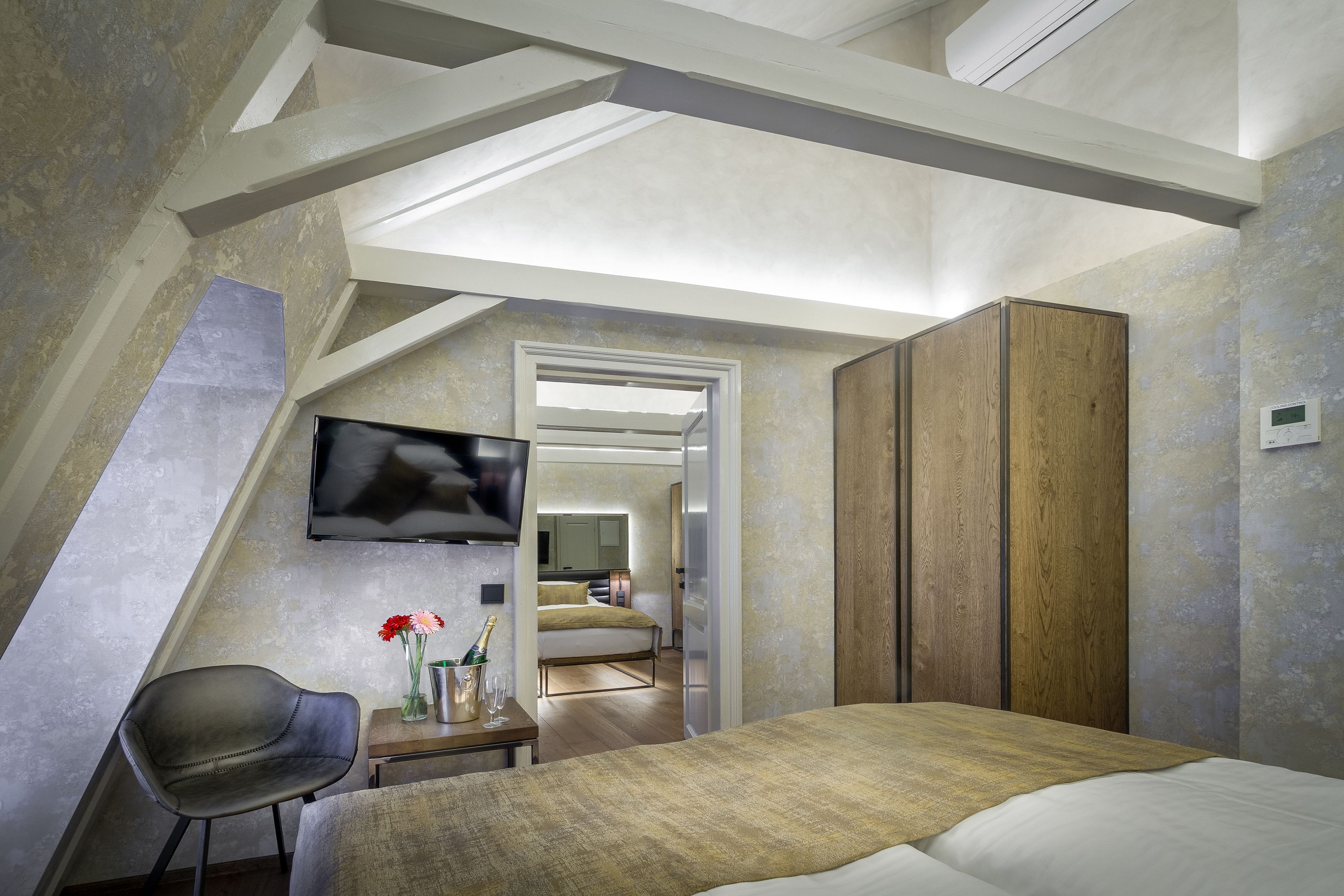 A bedroom with a sloped ceiling, a bed, a chair, a small table with a vase of flowers, and a television mounted on the wall.