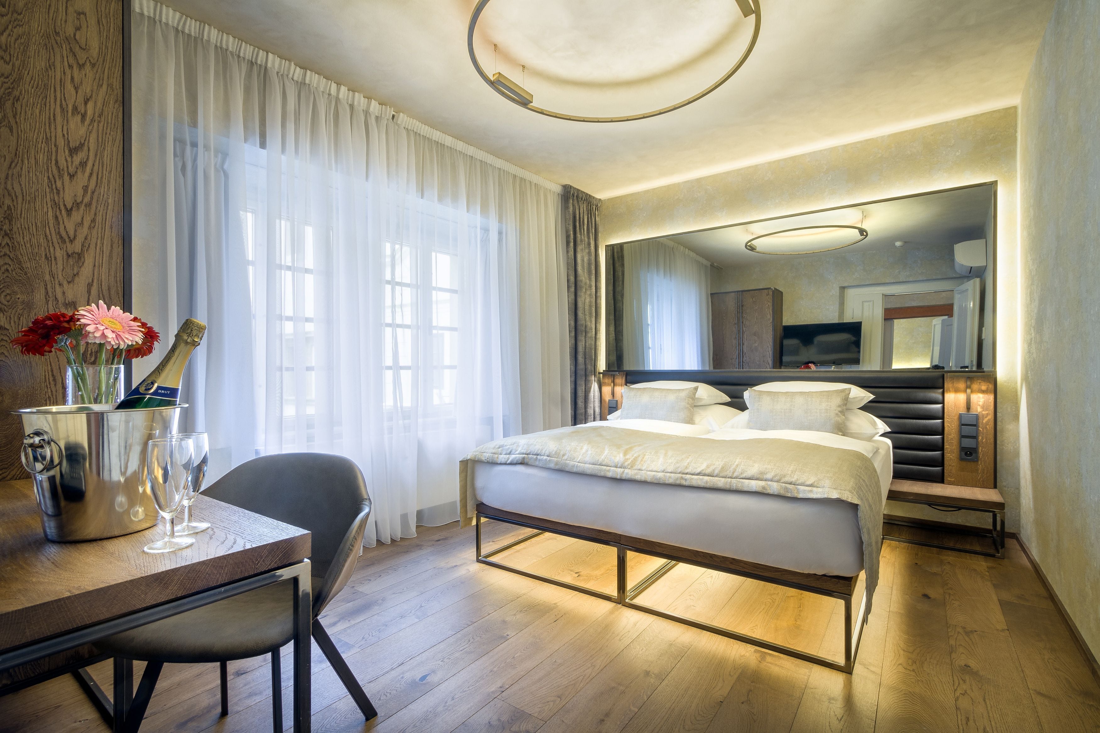 Ein modernes Hotelzimmer mit Holzboden, einem Bett mit Kopfteil, einem Schreibtisch mit Stuhl, einem großen Spiegel und einem Fenster mit durchsichtigen Vorhängen.