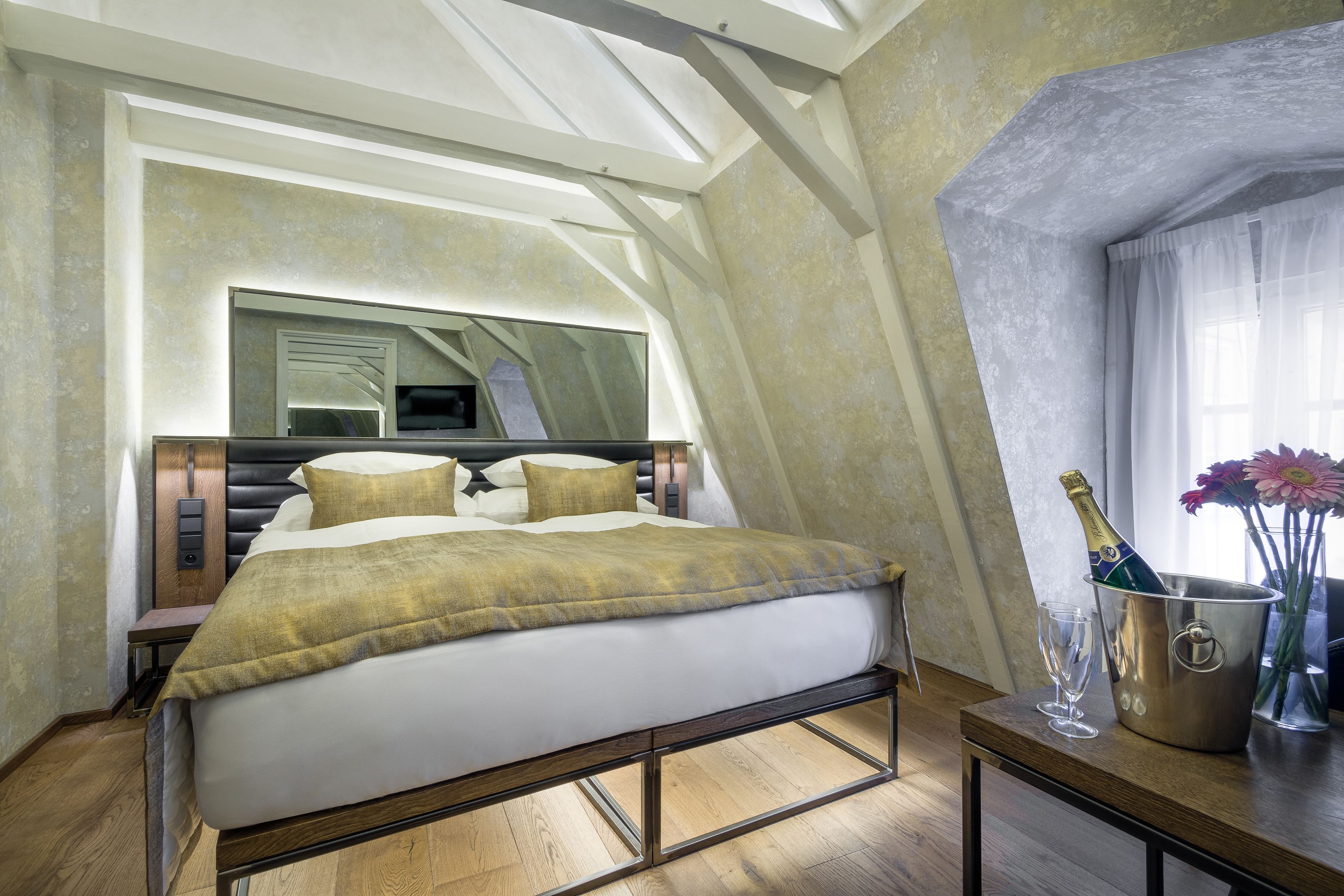 A modern bedroom with a large bed, a bedside table with a champagne bottle and glass, and a mirror above the bed.