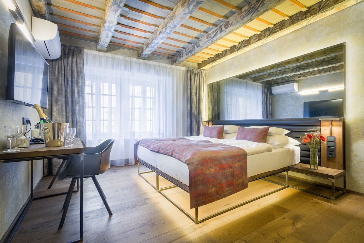A modern hotel room with a wooden ceiling, a large bed with a red blanket, a desk with a chair, and a TV.