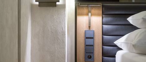 A modern hotel room with a bed, a control panel, and a small shelf.