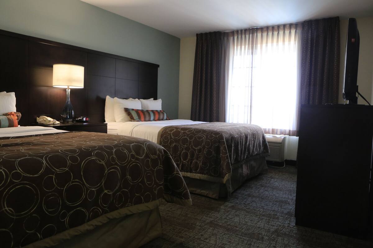 A hotel room with two beds, a nightstand with a lamp, a TV, and a large window with curtains.