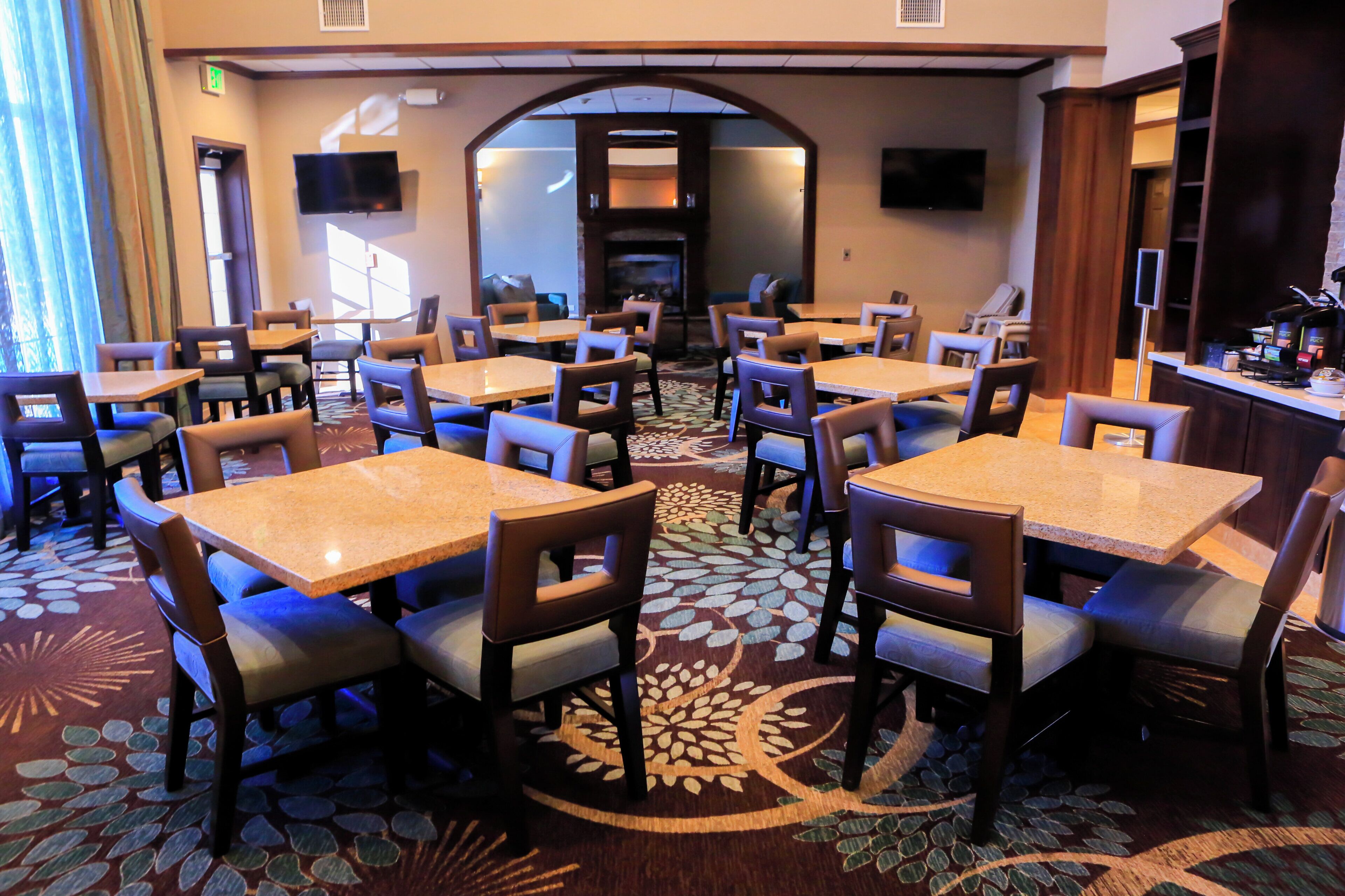 A hotel dining area with multiple tables and chairs, a patterned carpet, and a television screen.
