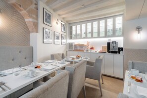 Un espace repas moderne avec des tables blanches, des chaises à revêtement gris et un bar pour le petit-déjeuner équipé d’une machine à café.