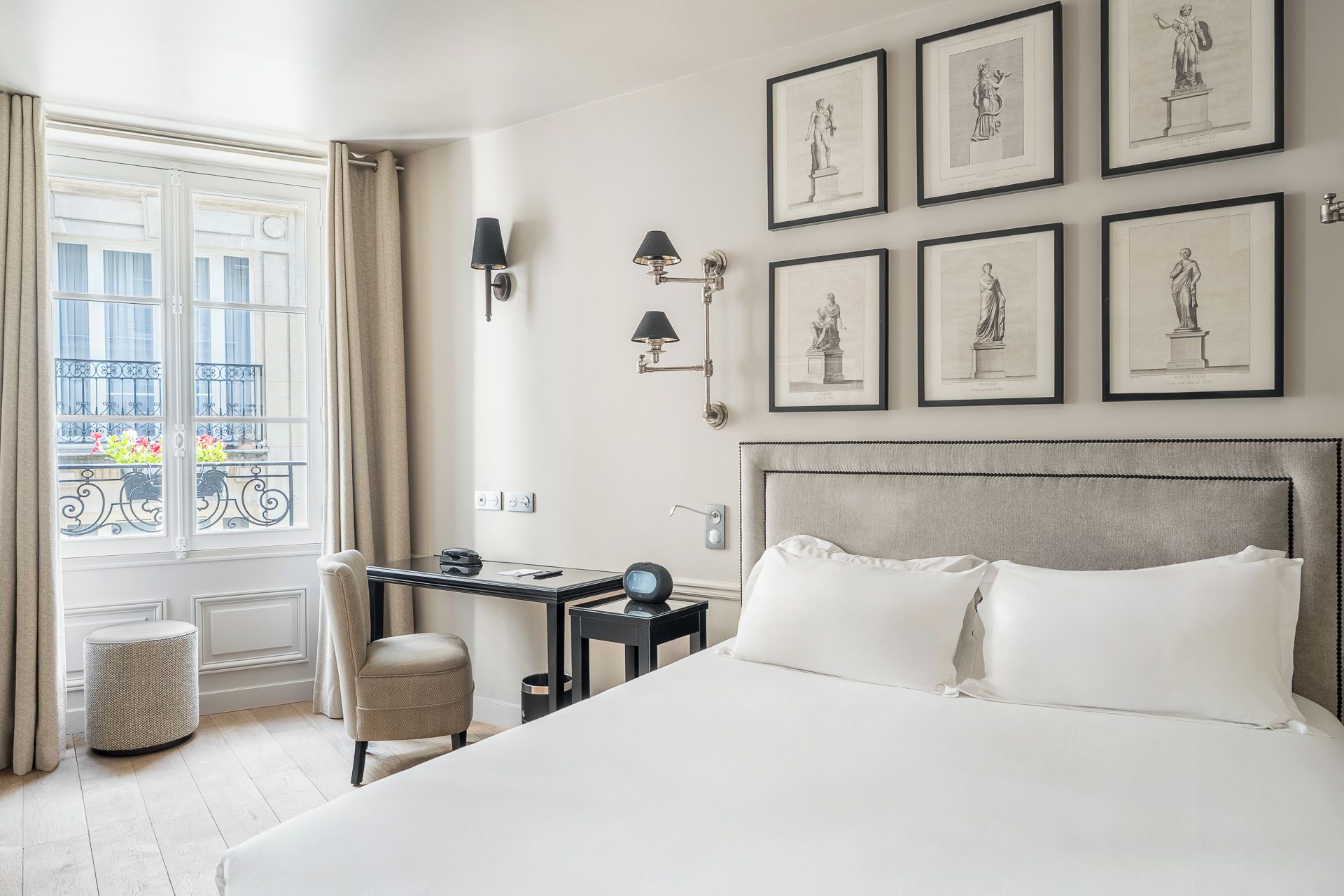 Habitación de hotel con una cama grande, un escritorio, una silla, un ventanal con cortinas y cuadros en la pared.