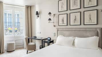 Habitación de hotel con una cama grande, un escritorio, una silla, un ventanal con cortinas y cuadros en la pared.
