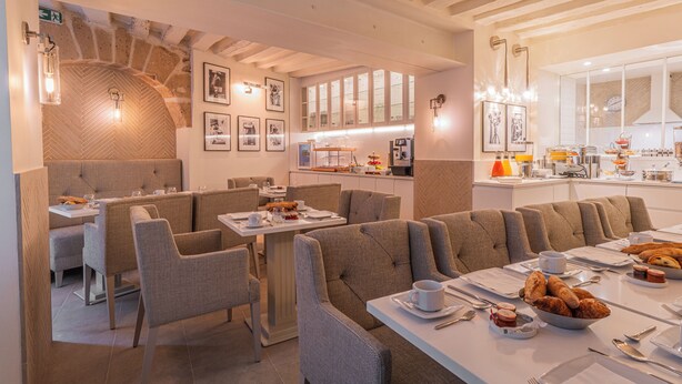 A modern dining area with high-backed chairs, a long communal table, and a kitchen in the background. - Hotel Résidence Des Arts - Saint-Germain (Paris)