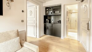 A modern kitchen with dark cabinets, a microwave, and a coffee maker. There is a wooden floor and a white door leading to a bathroom with a shower. - Hotel Résidence Des Arts - Saint-Germain (Paris)