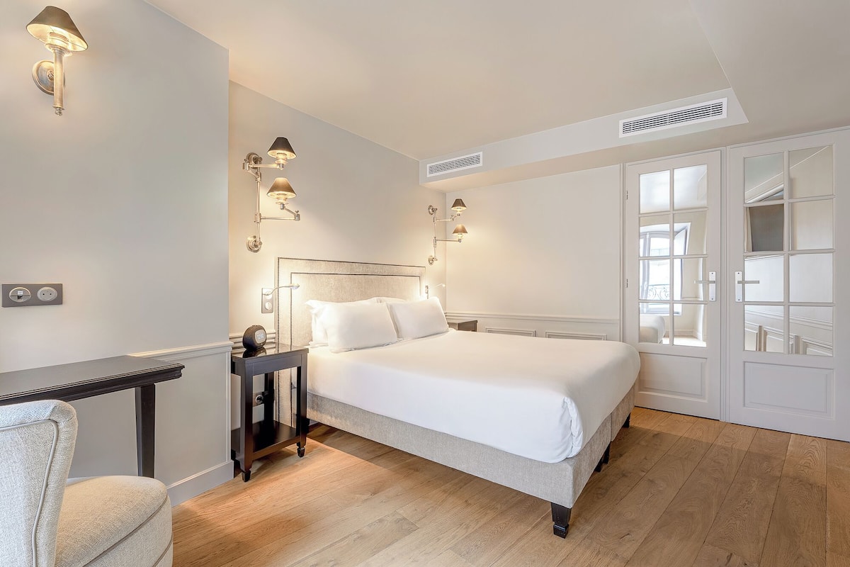 A bedroom with a bed, bedside tables, wall-mounted lamps, a nightstand, a chair, and a window with a view.