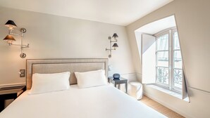 A hotel room with a large bed, two wall-mounted lamps, a small table with a clock, and a window with a view of the outside. - Hotel Résidence Des Arts - Saint-Germain (Paris)
