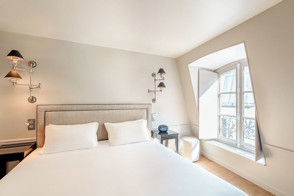 A hotel room with a large bed, two wall-mounted lamps, a small table with a clock, and a window with a view of the outside.