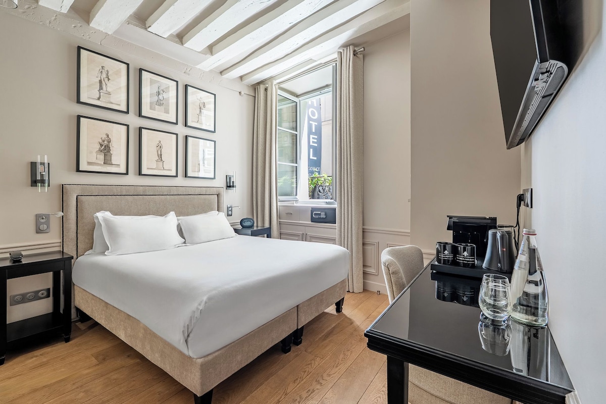 Una habitación de hotel con cama, mesitas de noche, escritorio con cafetera y cuadros en la pared.