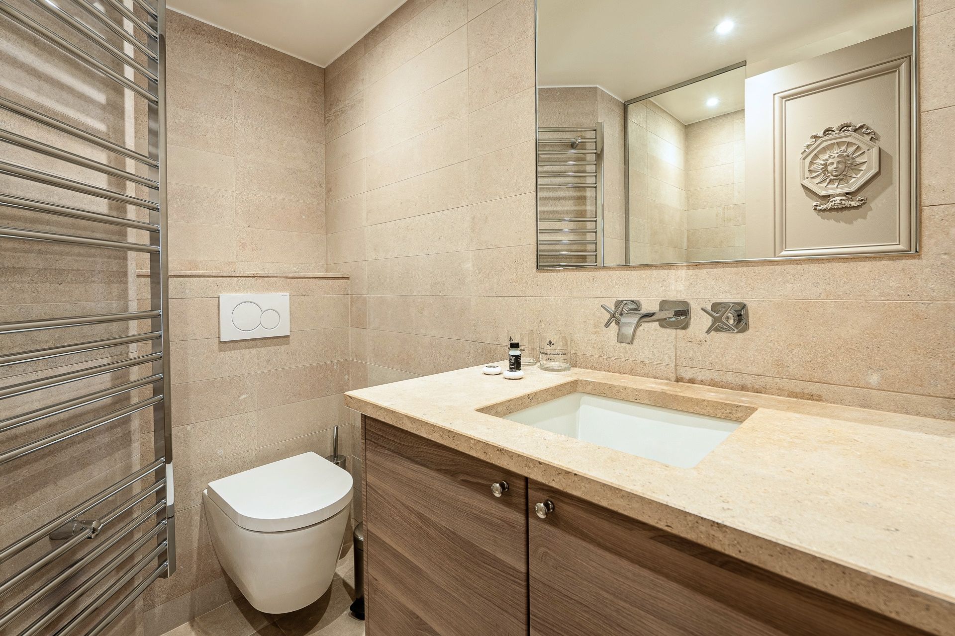A modern bathroom with a beige tiled wall, a white toilet, a wooden vanity with a sink, and a mirror with a decorative frame.