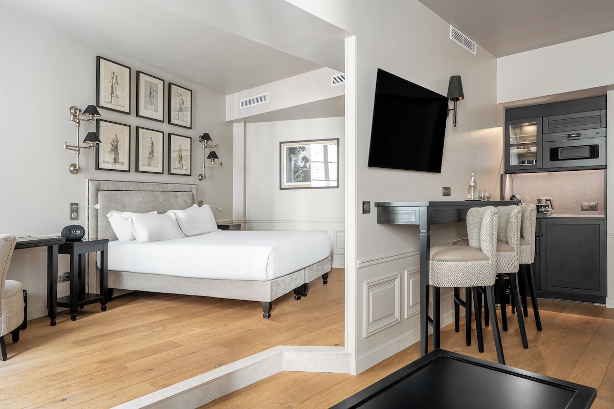Habitación de hotel con cama, mesita de noche, lámpara, cuadros enmarcados, televisor y kitchenette.