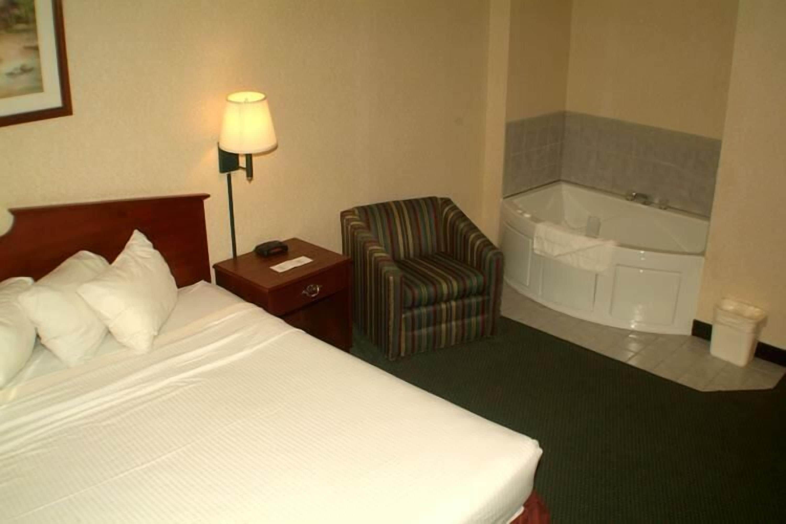 A hotel room with a bed, a chair, a nightstand, a lamp, and a bathroom with a bathtub.