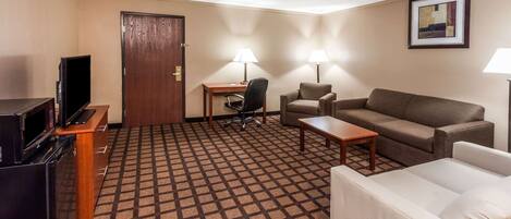 A hotel room with a sofa, a white armchair, a desk with a chair, a television, and a door.
