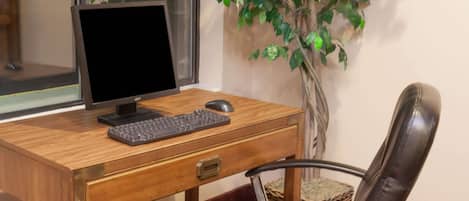 A wooden desk with a computer monitor, keyboard, and mouse. A black chair is in front of the desk. A potted plant is beside the desk.