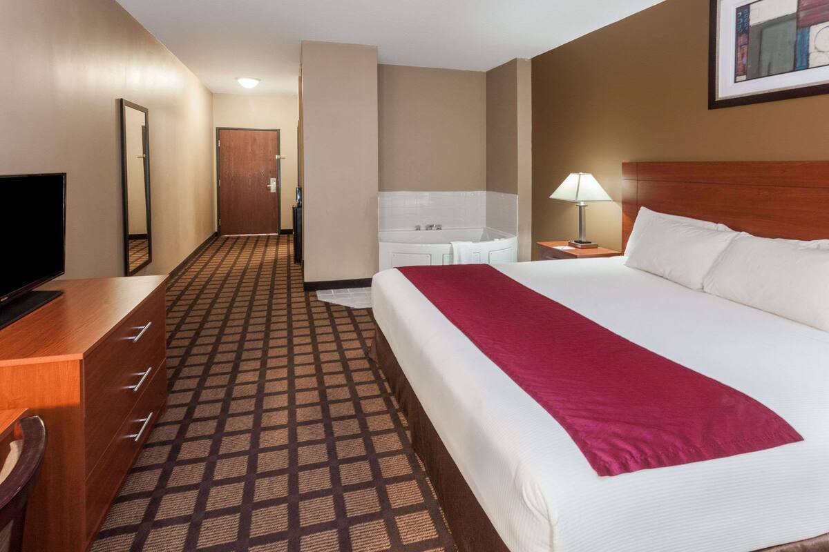 A hotel room with a large bed, a desk with a TV, a bathroom with a bathtub, and a patterned carpet.