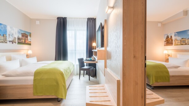 Een hotelkamer met twee bedden, een bureau en een stoel. Er hangen schilderijen aan de muren en een raam met gordijnen.