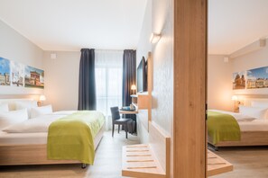 Une chambre d’hôtel avec deux lits, un bureau et une chaise. Les murs sont ornés de tableaux et il y a une fenêtre avec des rideaux.