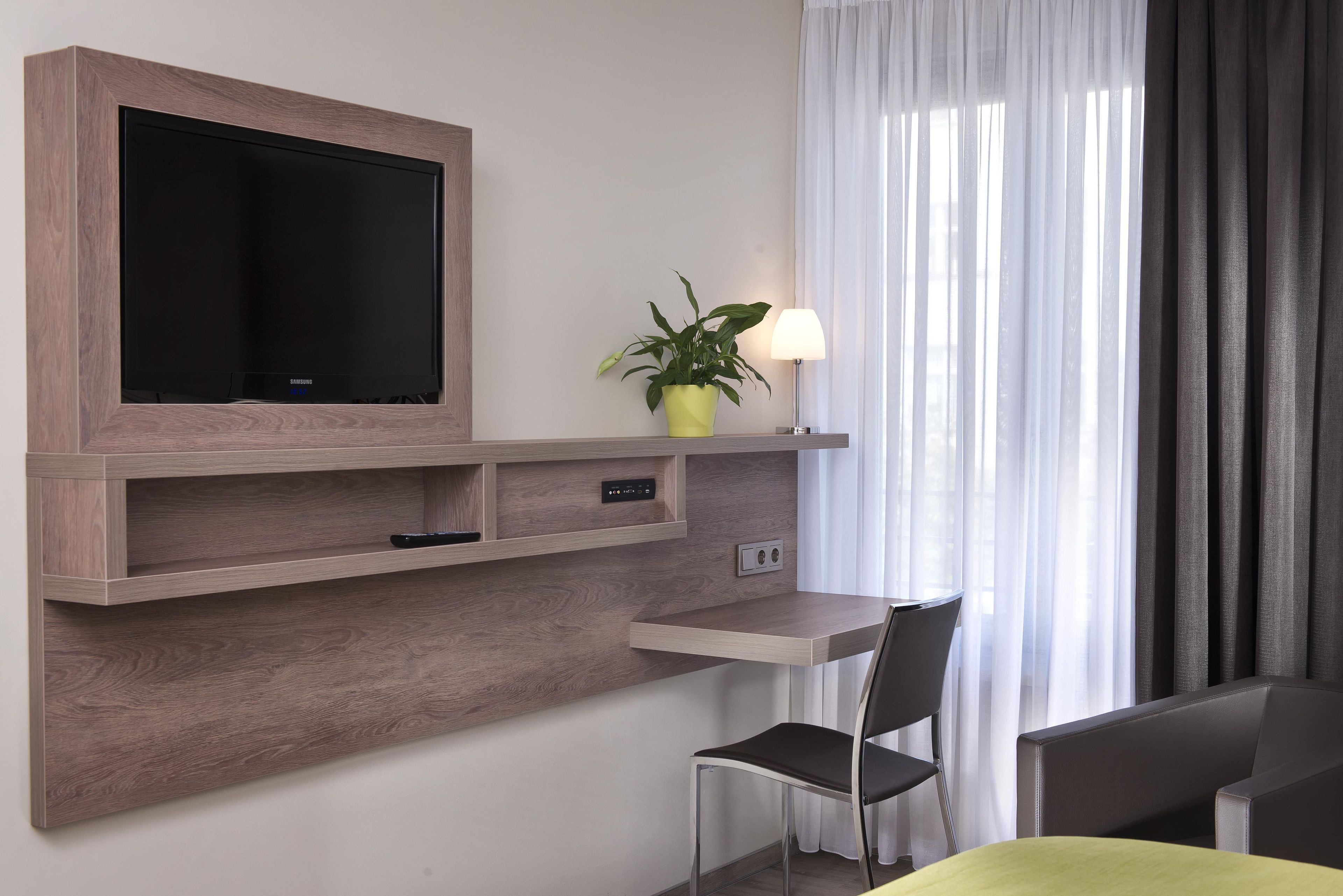 Une chambre moderne équipée d’un téléviseur fixé au mur, d’un bureau avec une chaise, d’une plante en pot et d’une lampe.