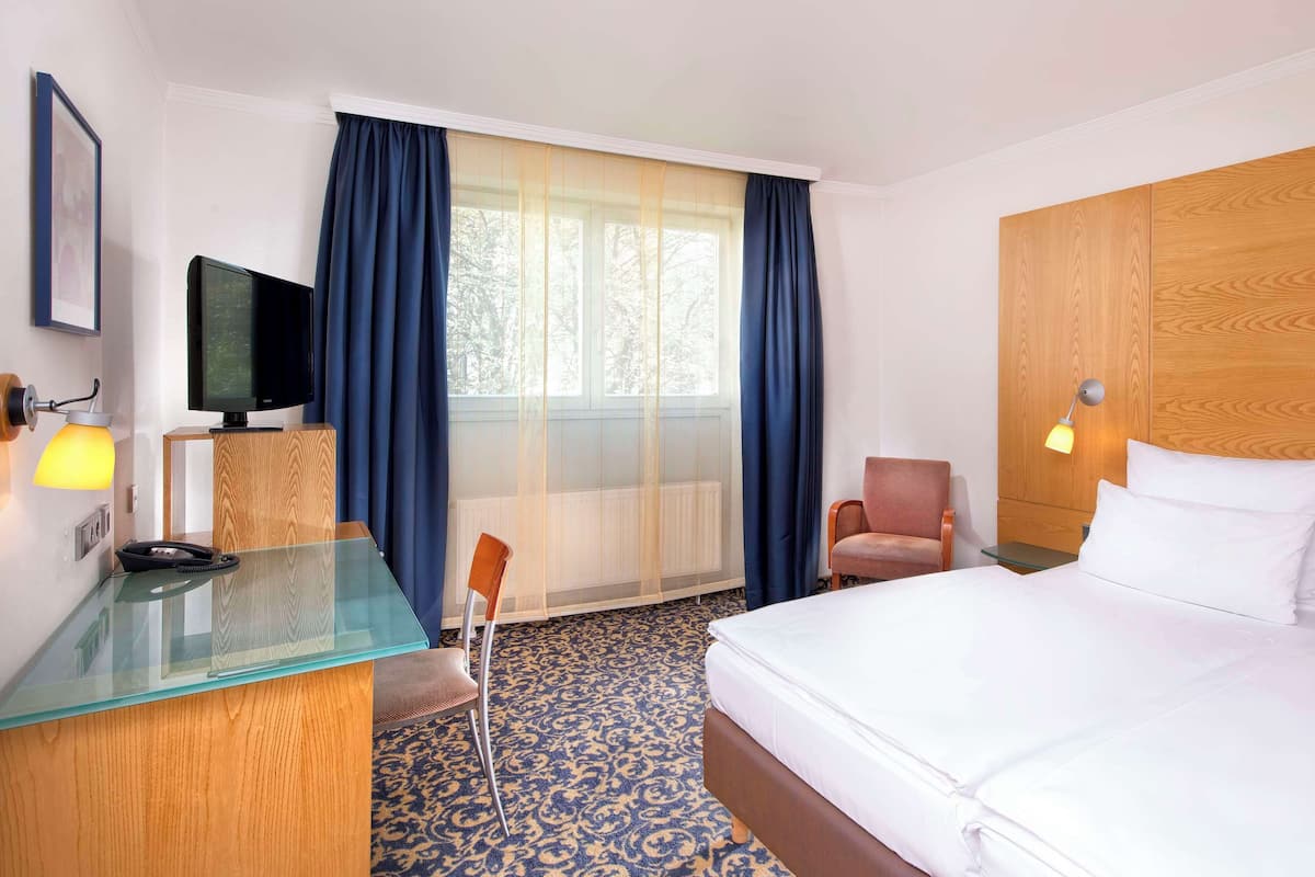 Ein Hotelzimmer mit einem Bett, einem Schreibtisch mit Computer, einem Sessel, einem Fernseher und einem Fenster mit Vorhängen.