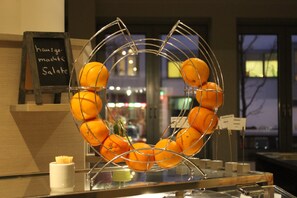 Un présentoir métallique rempli d’oranges, un panneau d’ardoise et une vue sur la rue à travers la fenêtre.