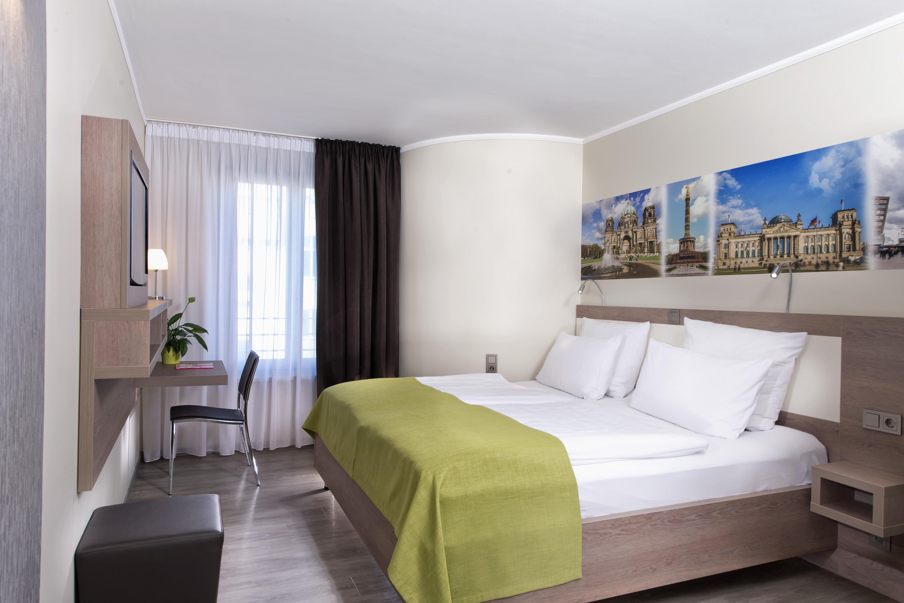 A hotel room with a bed, a desk, a chair, a plant, and a large window with curtains.