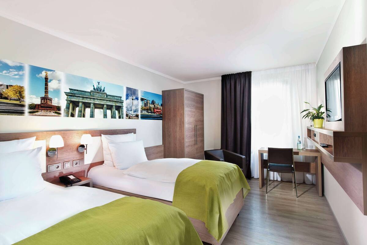 Ein Hotelzimmer mit zwei Betten, einem Schreibtisch, einem Stuhl und einem großen Fenster mit Vorhängen.