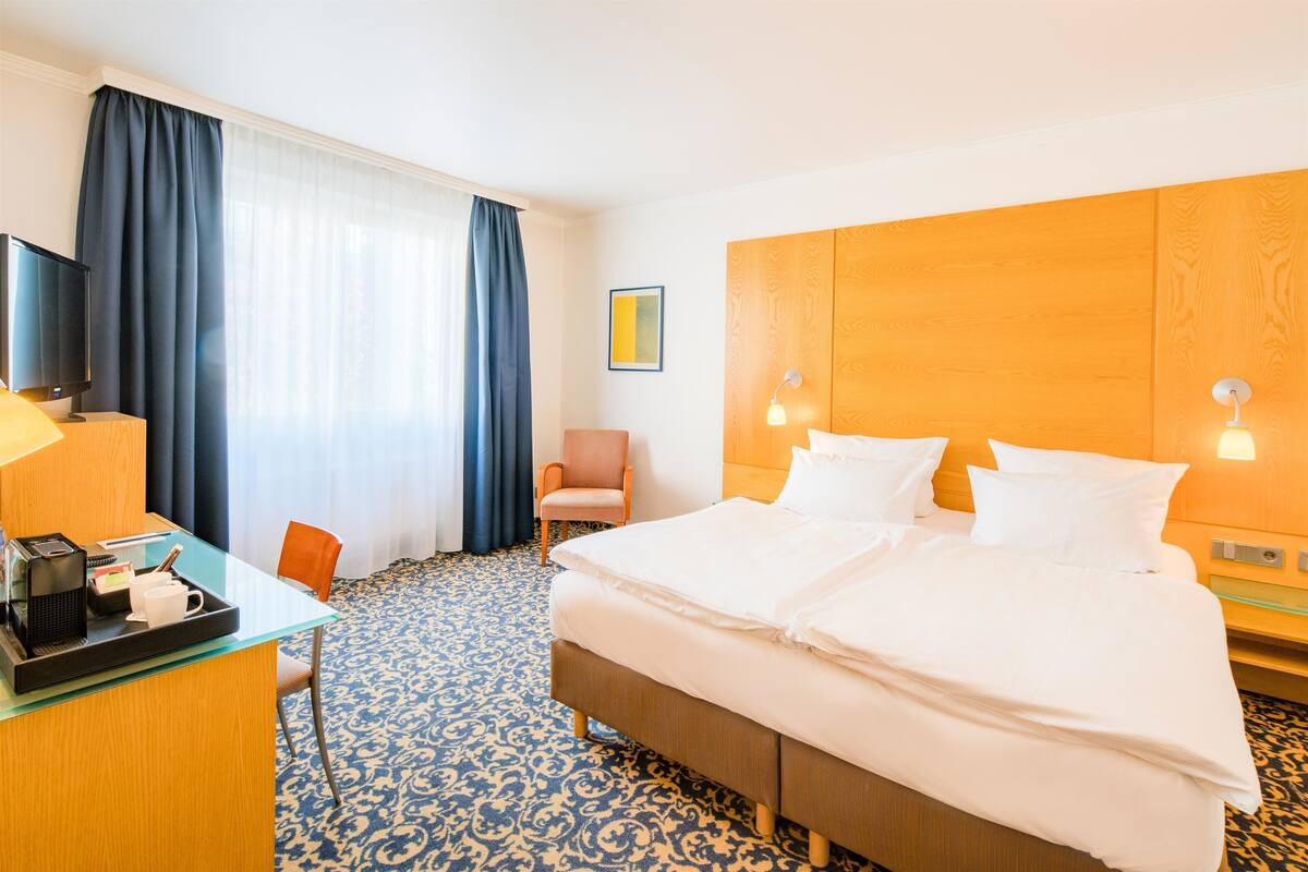 Ein Hotelzimmer mit einem Bett, einem Schreibtisch mit Kaffeemaschine, einem Sessel, einem Fernseher und einem fenster mit Vorhängen.