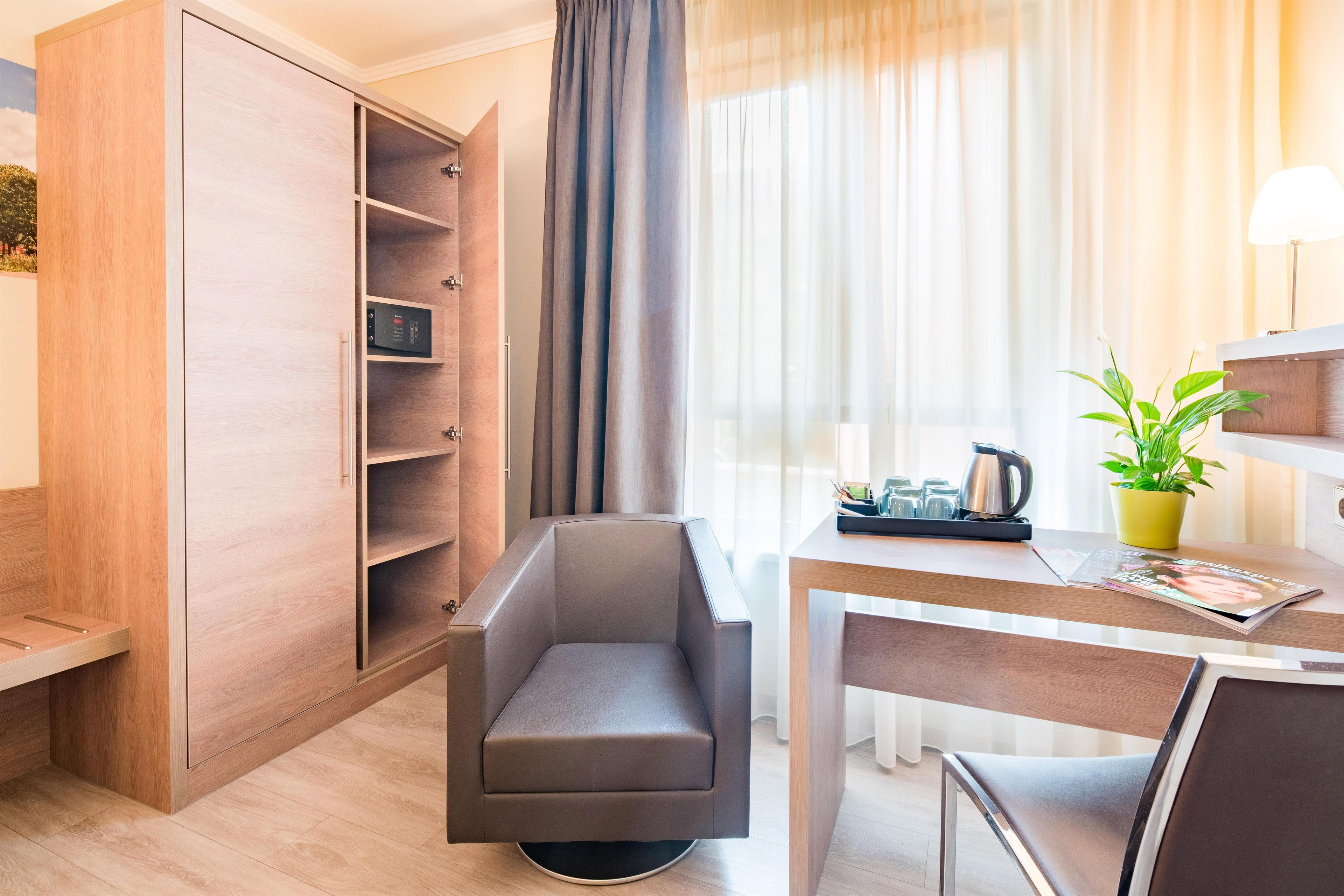 Moderni hotellihuone, jossa on puinen vaatekaappi, harmaa nojatuoli, ruokapöytä, jossa on lehti, kahvinkeitin ja ruukkukasvi.