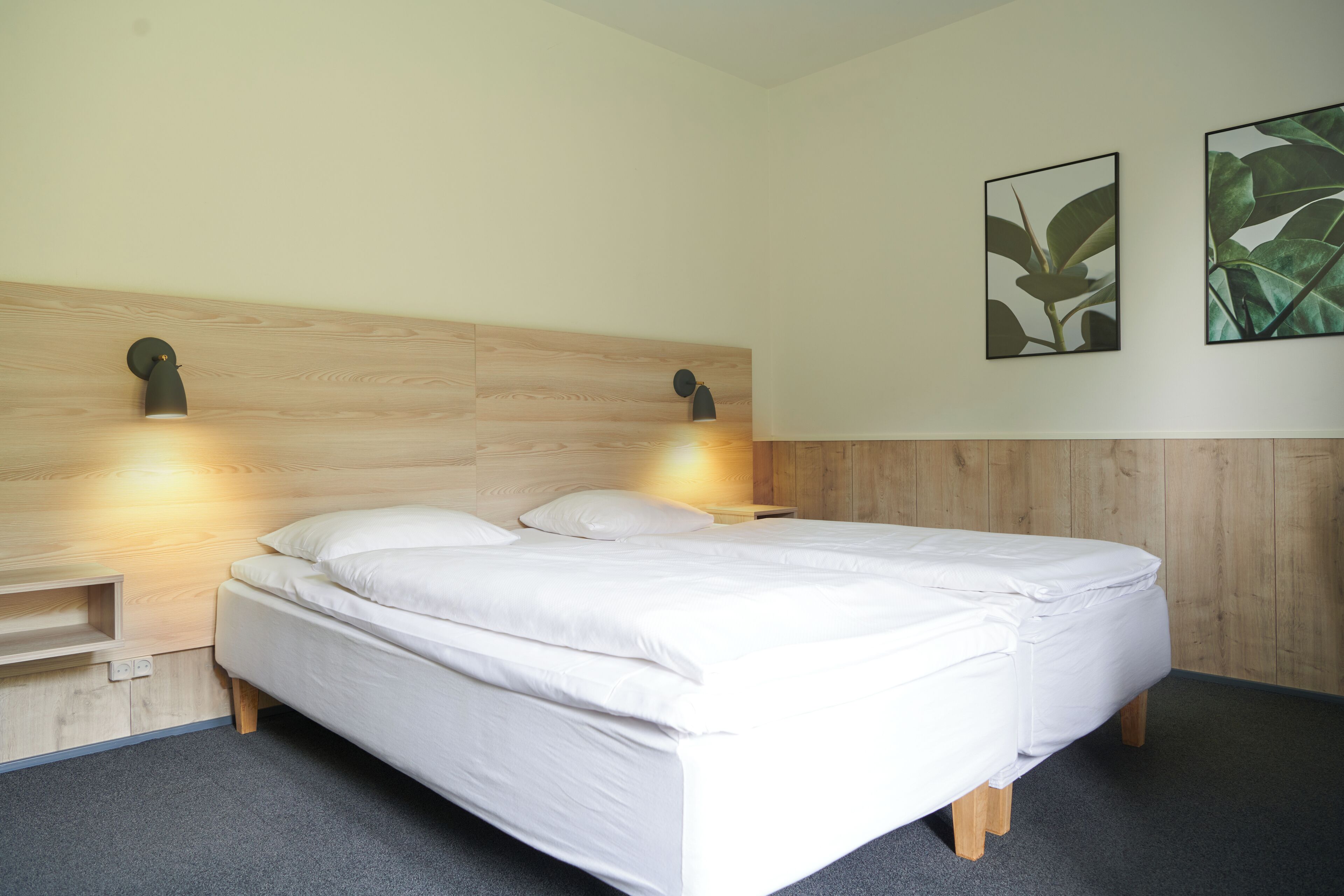 A neatly made bed with a wooden headboard, two wall-mounted lights, and two framed botanical prints on the wall.
