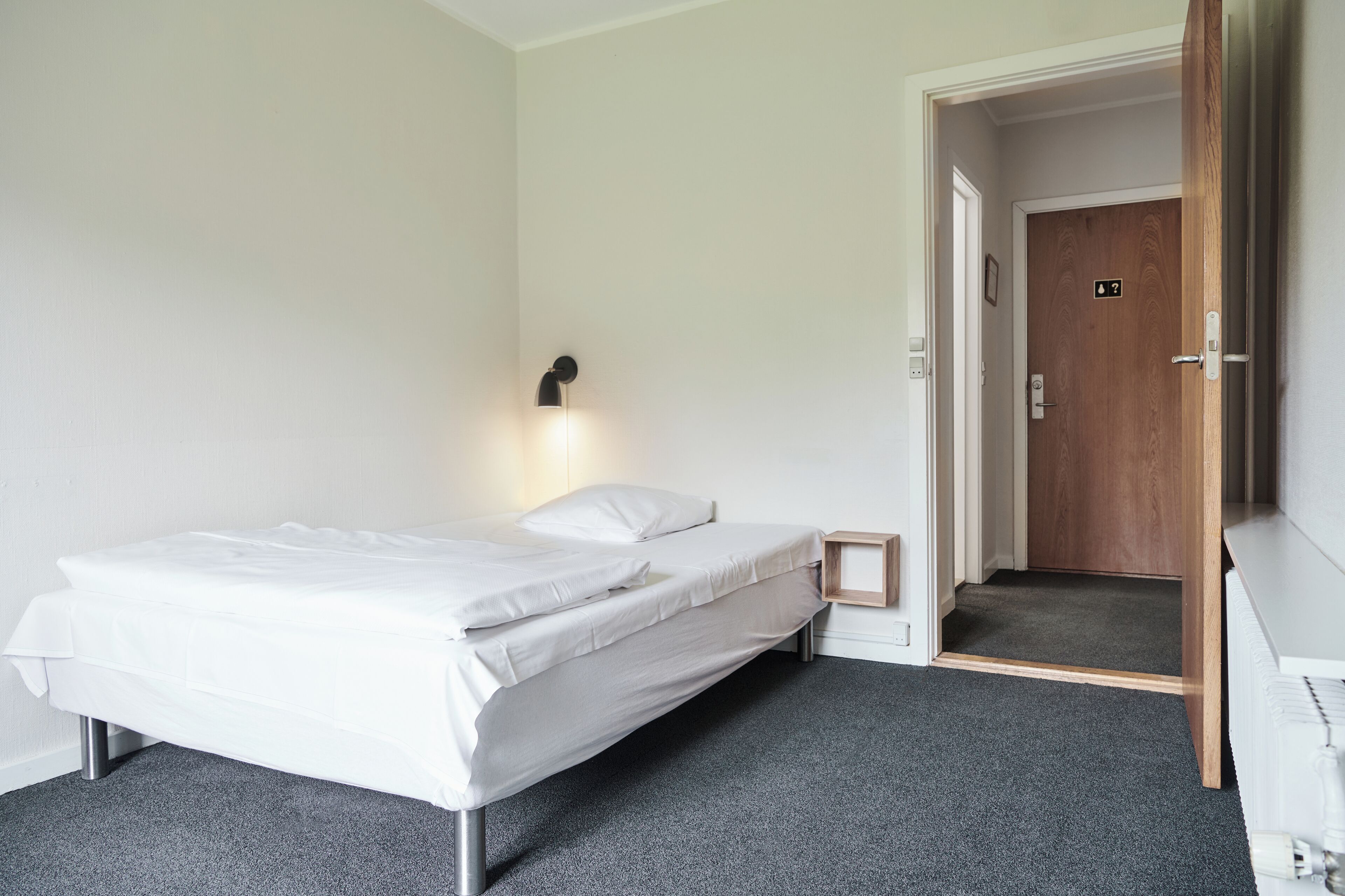 Un lit simple avec un matelas et des oreillers blancs, une petite table de chevet, une lampe fixée au mur et une porte donnant sur un couloir.