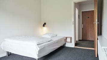 Un lit simple avec un matelas et des oreillers blancs, une petite table de chevet, une lampe fixée au mur et une porte donnant sur un couloir.