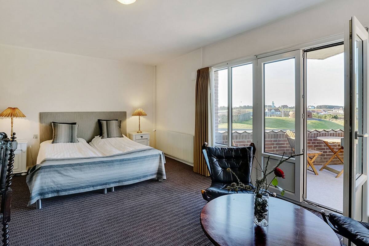 Habitación de hotel con cama, dos lámparas de noche, una silla, una mesa y un ventanal con vistas al exterior.