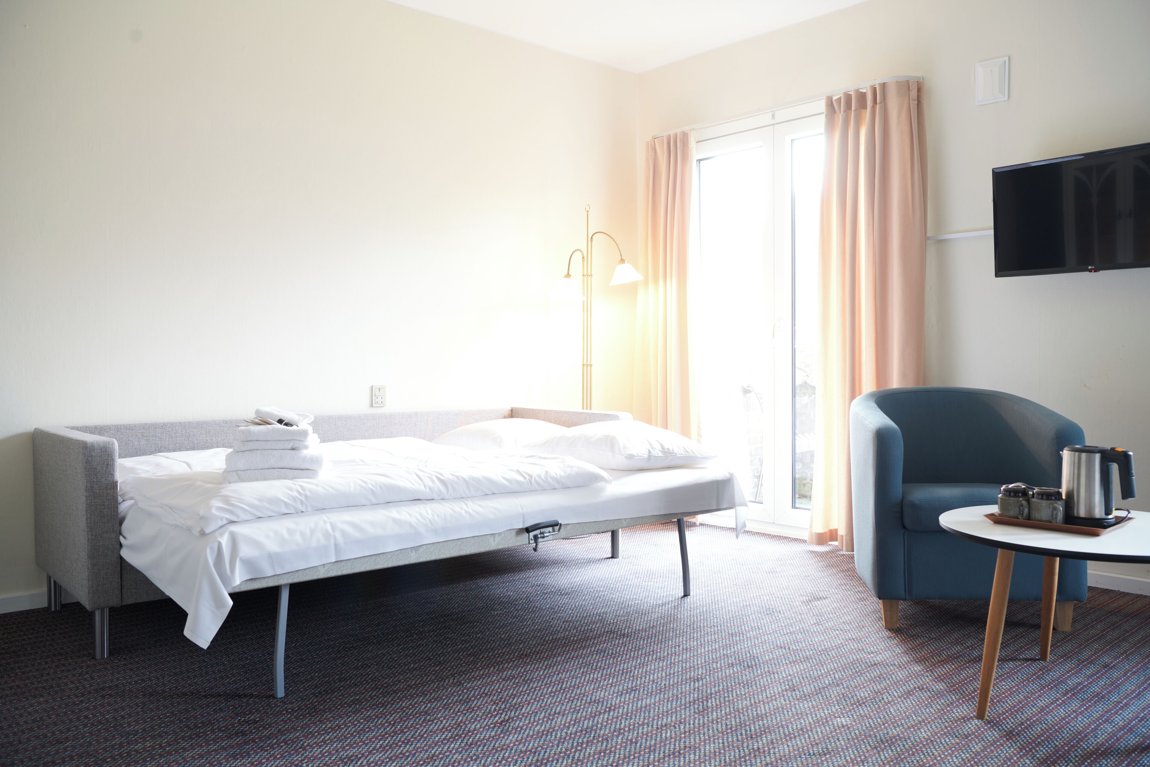 A hotel room with a bed, a chair, a table, a TV, and a lamp.